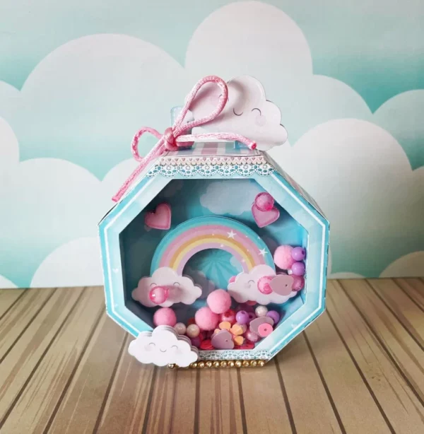 Embalagem vertical estilo caixa de leite na cor rosa pastel com estampa de mini nuvens brancas, destacando na parte frontal um grande aplique de arco-íris colorido em camadas tridimensionais em arquivo digital de corte.