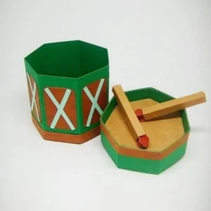 Caixinha de papel estruturada em formato cilíndrico imitando um tambor musical com baquetas cruzadas aplicadas no topo, detalhes de cordas cruzadas nas laterais e bordas coloridas em camadas tridimensionais em arquivo digital de corte.