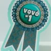 Topper de bolo com um brasão de super-herói estilizado com a letra V no centro, cercado por raios brilhantes amarelos, uma capa esvoaçante azul e estrelas em camadas tridimensionais em arquivo digital de corte.