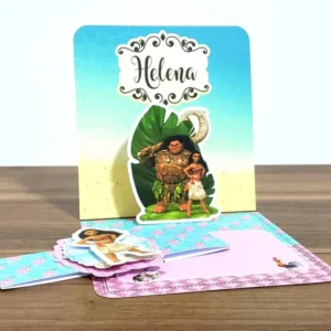 Convite impresso aberto exibindo o efeito pop up tridimensional onde a personagem Moana surge em pé na proa de seu barco a vela sobre ondas do mar esculpidas em papel azul, com o porco Pua e o galo Heihei na areia da praia, em arquivo digital de corte.