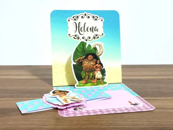 Convite impresso aberto exibindo o efeito pop up tridimensional onde a personagem Moana surge em pé na proa de seu barco a vela sobre ondas do mar esculpidas em papel azul, com o porco Pua e o galo Heihei na areia da praia, em arquivo digital de corte.