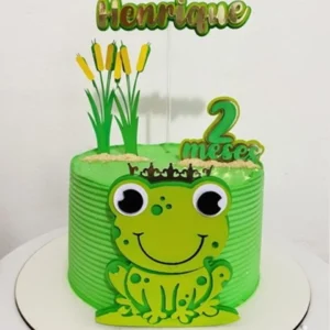 Topper de bolo com um sapinho verde fofo e sorridente sentado sobre uma vitória-régia redonda, cercado por pequenas flores aquáticas e taboas em camadas tridimensionais para decoração de festa em arquivo digital de corte.