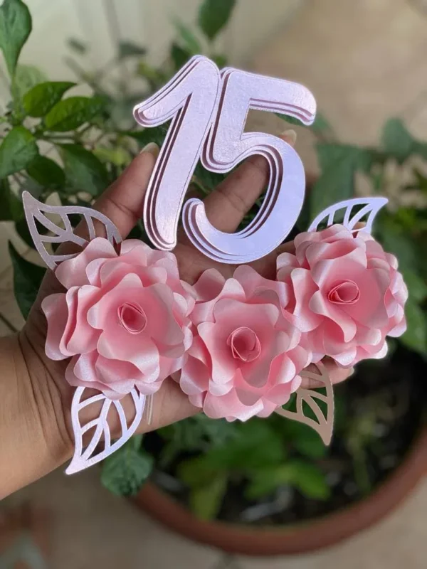 Topper de bolo de debutante com o número 15 recortado em letras elegantes com brilho dourado, cercado por arranjos de rosas abertas em tons de rosa e delicados ramos de folhagens dispostos em camadas tridimensionais em arquivo digital de corte.
