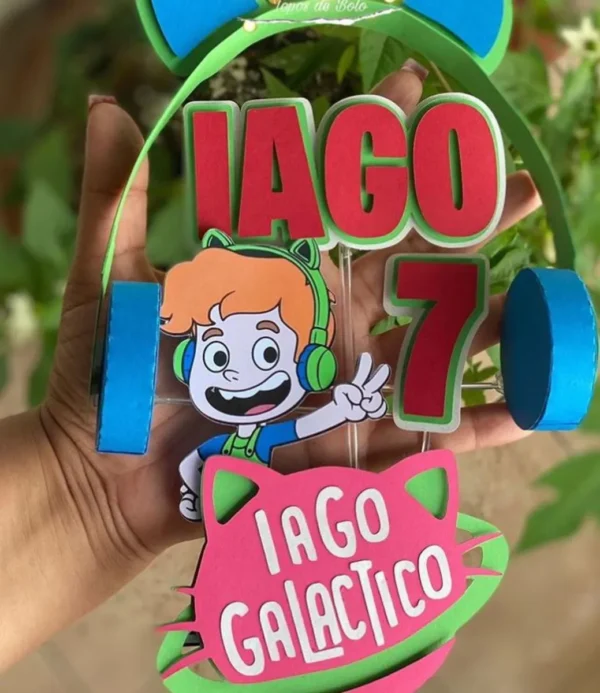 Topper de bolo em camadas com o tema do Gato Galáctico, exibindo o personagem de capuz azul, estrelas sorridentes, foguetes coloridos e uma faixa central amarela para nome.
