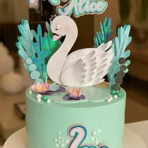 Topper de bolo com um cisne branco majestoso usando uma coroa dourada brilhante, cercado por flores em tons de rosa claro e pêssego, ramos de folhas verdes delicadas e detalhes em glitter dourado em camadas tridimensionais em arquivo digital de corte.