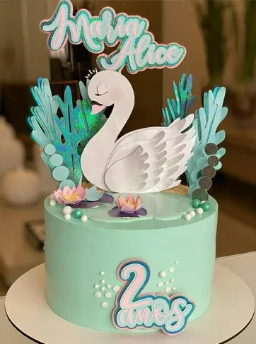 Topper de bolo com um cisne branco majestoso usando uma coroa dourada brilhante, cercado por flores em tons de rosa claro e pêssego, ramos de folhas verdes delicadas e detalhes em glitter dourado em camadas tridimensionais em arquivo digital de corte.