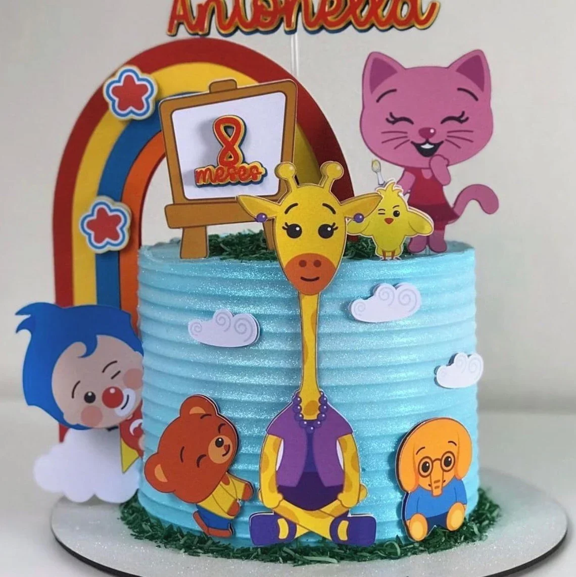 Topper de bolo com o palhacinho Plim Plim sorridente de braços abertos, usando sua roupa vermelha com uma grande estrela amarela no peito, cercado por um arco-íris colorido, nuvens brancas e notas musicais em camadas tridimensionais em arquivo digital de corte.